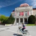 Blick auf die Oper von Graz. Im Vordergrund fährt eine junge Frau mit dem Fahrrad.